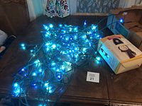 Tangled blue Christmas string lights illuminated on a dark surface, next to a box with a snowman image, labeled with lot number 21.