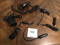 Six black charging cords on wooden surface, five with cigarette lighter plugs and one with wall plug adapter.