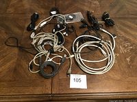 Photo showing a collection of assorted electronic cords and cables arranged on a wooden surface, predominantly black and white, loosely coiled and bundled.