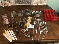 Photo showing various cutlery including knives, forks, spoons, disposable plastic utensils, wooden chopsticks, and a red plastic utensil organizer on a wooden table.