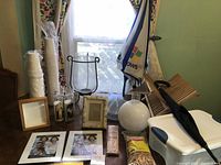 Photo showing glass hurricane lamp with black metal stand, religious glass candles, stacked white disposable cups, assorted picture frames, cooler, umbrella, and other items on table near window.