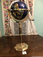 Full view of the large world globe on brass stand placed on a wooden surface with floral curtains in background.