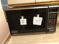 Front view of the black microwave on countertop showing the entire unit with two stickers on the door indicating '72' and '24 x 14' dimensions.