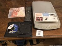 HP PhotoSmart flatbed scanner with open CD tray showing included software CD, power adapter, and part of installation guide booklet on a wood table.