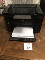 Black HP LaserJet P1606dn printer with paper output tray holding paper on a wooden surface.