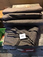 Photo showing four thick, dark gray wool blankets neatly folded and stacked on a metal surface with a lot number card "108" placed on top.