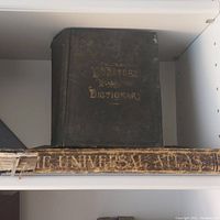 Photo showing Webster's Unabridged Dictionary with black cover and gold embossed title, placed above The Universal Atlas with damaged spine