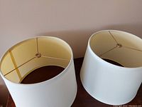 Photo of two cream colored cylindrical lampshades showing interior wire frame and top fitter, placed side by side on a wood surface against a beige wall.