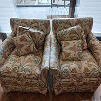 Pair of small armchairs upholstered in lotus pattern fabric with four matching cushions, set near a large window, providing an overall view of both chairs.