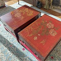 Pair of rectangular red lacquer Chinese trunk-form coffee tables on stands, top view showing floral decorations, one table with yellow flowers and the other with pink flowers and a bird, sitting on a patterned rug in a living room.