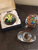 Three glass paperweights displayed together, including Caithness 'Flower in the Rain' in box, multicolor floral, and clear glass with eye form