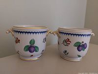 Two white porcelain cache pots with colorful fruit and floral hand-painted decorations and gold accents, displayed side by side on a light surface.