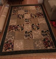 Full view of area rug on hardwood floor showing patchwork squares, floral and textured pattern, with dark border.