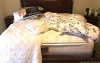 Full bed showing mattress with white textured blanket, floral patterned comforter, and a stack of pillows and linens on the side.