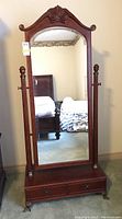 Full view of the cheval mirror showing carved top and side finials, lower drawer, and reflection of room.