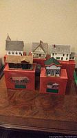 Five miniature buildings representing a Christmas village set displayed on red boxes.