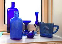 Seven pieces of vintage cobalt blue glass arranged on a white surface in front of a window. Includes various bottles, pedestal dish, chicken bowl, vase, and pitcher.