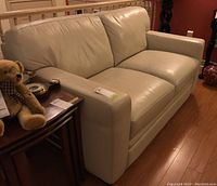 Side angle showing cream leather love seat with padded armrest and some staining visible on armrest.
