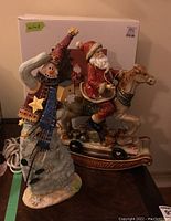 Photo of ceramic Santa Claus figurine seated on a rocking horse with original box behind, next to a tall snowman figurine.