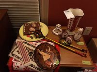 Photo showing many items together on two tabletops including plates, cloth runners, wooden spoons, figurine, eggs, vase, and candlestick.