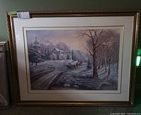 Framed winter landscape print showing a large cozy cabin in snow at sunrise with horse and sleigh in foreground, deep gold ornate frame.