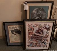Three framed vintage prints leaning against the wall, showing a variety of artistic and historical designs including music sheets and a cigar advertisement poster