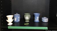 Five Wedgwood jasperware pieces displayed on a black shelf, including blue, green, and white vases and a covered box with decorative white reliefs