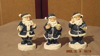 Photo shows three vintage Santa Claus figurines wearing blue coats and white fur trim. Each holds different small holiday items and stands on a white base.