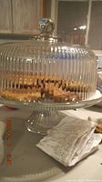 Clear glass cake stand with ribbed dome lid and round knob on top, shown on counter with crumpled receipts nearby.