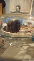 Glass pedestal cake stand with dome cover holding a partially eaten chocolate cake.