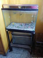Large fish aquarium on metal stand with light fixture, containing gravel and glass fish ornaments. Smaller empty fish tank underneath on shelf.