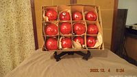 Top view of a cardboard box containing 12 round red glass Christmas ornaments arranged in four rows of three. Each ornament is shiny red with a metal hanging cap.