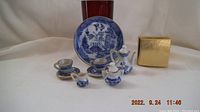 Miniature Japanese tea set arranged with cups, teapot, creamer, sugar bowl, plate and a gold-colored box on a white background.