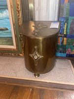 Front view of curved brass kindling box showing decorative design on front and lid, brass side handles, and patina on surface.