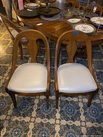 Pair of wooden dining chairs with cushioned white seats side by side in front of a wooden dining table.