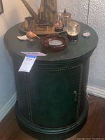 Full view of green round end table cabinet from front, showing its door, decorative beaded trim, and top surface with various objects.