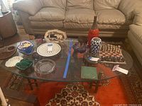 Wide angle photo showing the glass coffee table with various objects placed on top, including decorative pottery, chess set, and vases on an orange and patterned rug with a beige leather sofa in background.