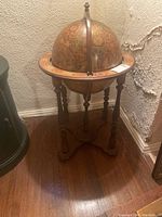 Full view of the vintage globe bar cart standing on wooden floor against textured wall, showing overall shape and condition with missing foot/peg on one leg.