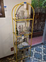 Photo of one yellow metal 4-tier arched shelf unit displaying various glassware and ceramics on each shelf.
