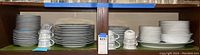 Wide view of the stacked dinner plates, salad plates, dessert plates, tea cups and saucers on wooden shelf showing quantity and pattern.