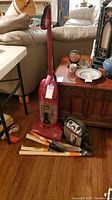 Red Eureka WideTrack SuperLite upright vacuum cleaner beside a Vector jump starter pack and several gardening tools on a wood floor in a living room area.