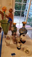 Group of seven vases and three blown glass flowers of various sizes arranged on a white table, showing Asian-themed floral patterns, some with wooden stands.