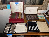 Four fitted cutlery cases on table including black case with silver spoons, black case with mother-of-pearl handled knives, white Hors d’Oeuvres Master box with forks, red case with steak knives