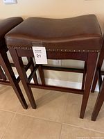 Close-up of one bar stool showing brown faux leather seat with nailhead trim and wooden legs.