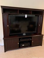 Front view of dark cherry wood entertainment center with a 55 inch TV placed inside, showing vertical slatted doors and silver handles.