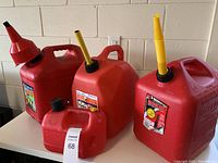 Front and side view of four red plastic gas cans arranged in a group: three large 5-gallon cans and one smaller 1-gallon can, displayed on a white surface against a light wall background.