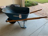 Side view of Jackson steel wheelbarrow showing blue metal tray, wooden handles, black rubber wheel, and support legs.