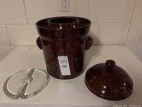 Brown ceramic pickling crock with handles, matching lid and two glass weights visible beside it on white countertop.