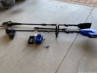 Overview of two long shafts with saw and edger heads plus battery and charger on concrete floor