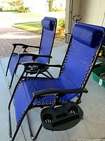 Two blue reclining camping chairs shown side by side, highlighting adjustable headrests and black side trays with cup holders attached to the armrests.